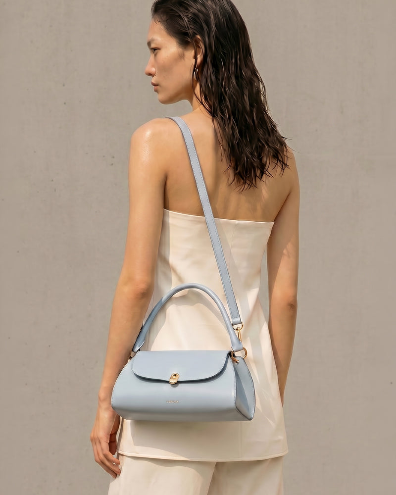 A woman shown from the back wearing a baby blue leather shoulder bag with a top handle and gold-tone hardware, paired with a cream strapless top against a neutral gray background.