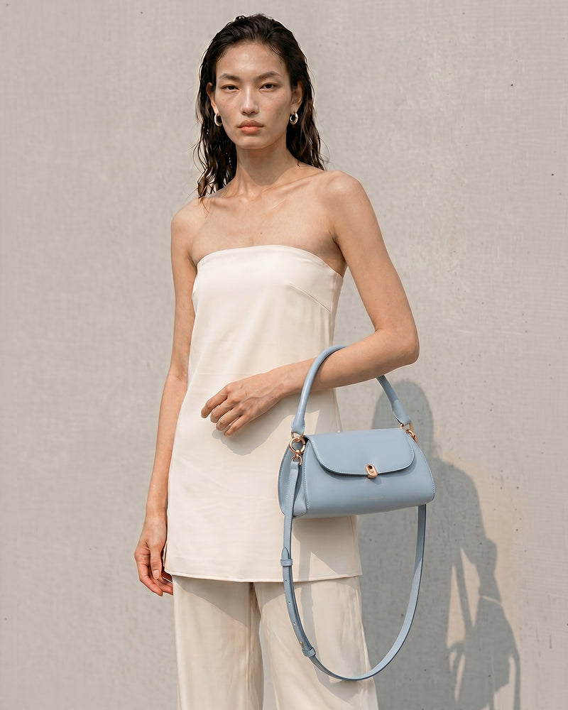 A model wearing a white strapless top and matching trousers holds a light baby blue leather shoulder bag with a matching strap against a textured light gray wall.