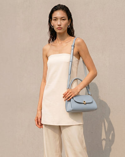 A model wearing a cream strapless top and beige trousers carries a light baby blue leather shoulder bag with a top handle and a thin matching shoulder strap against a textured beige wall.