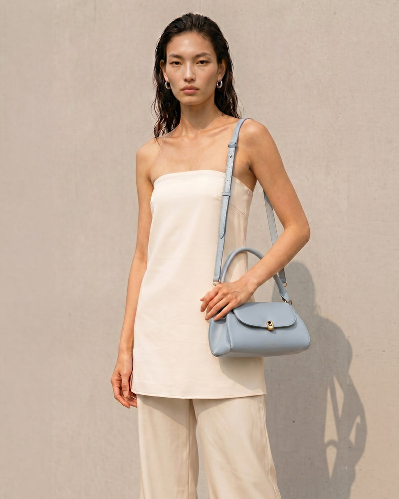 A model wearing a cream strapless top and beige trousers carries a light baby blue leather shoulder bag with a top handle and a thin matching shoulder strap against a textured beige wall.