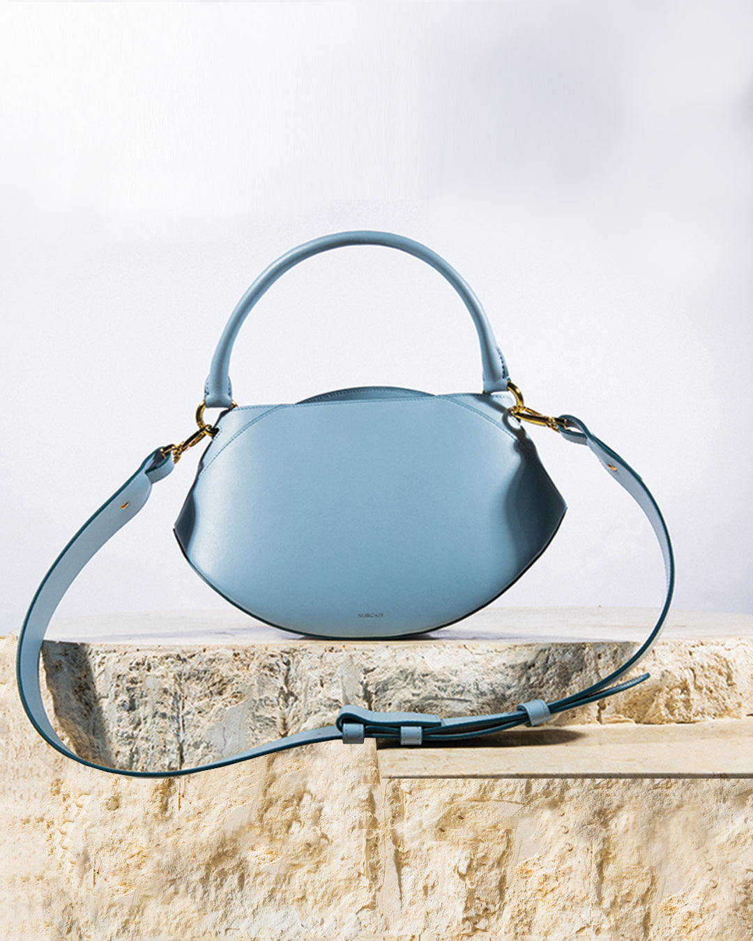 Sunrise Bag in Baby Blue shown from a three-quarter angle with its wide leather shoulder strap on a white background
