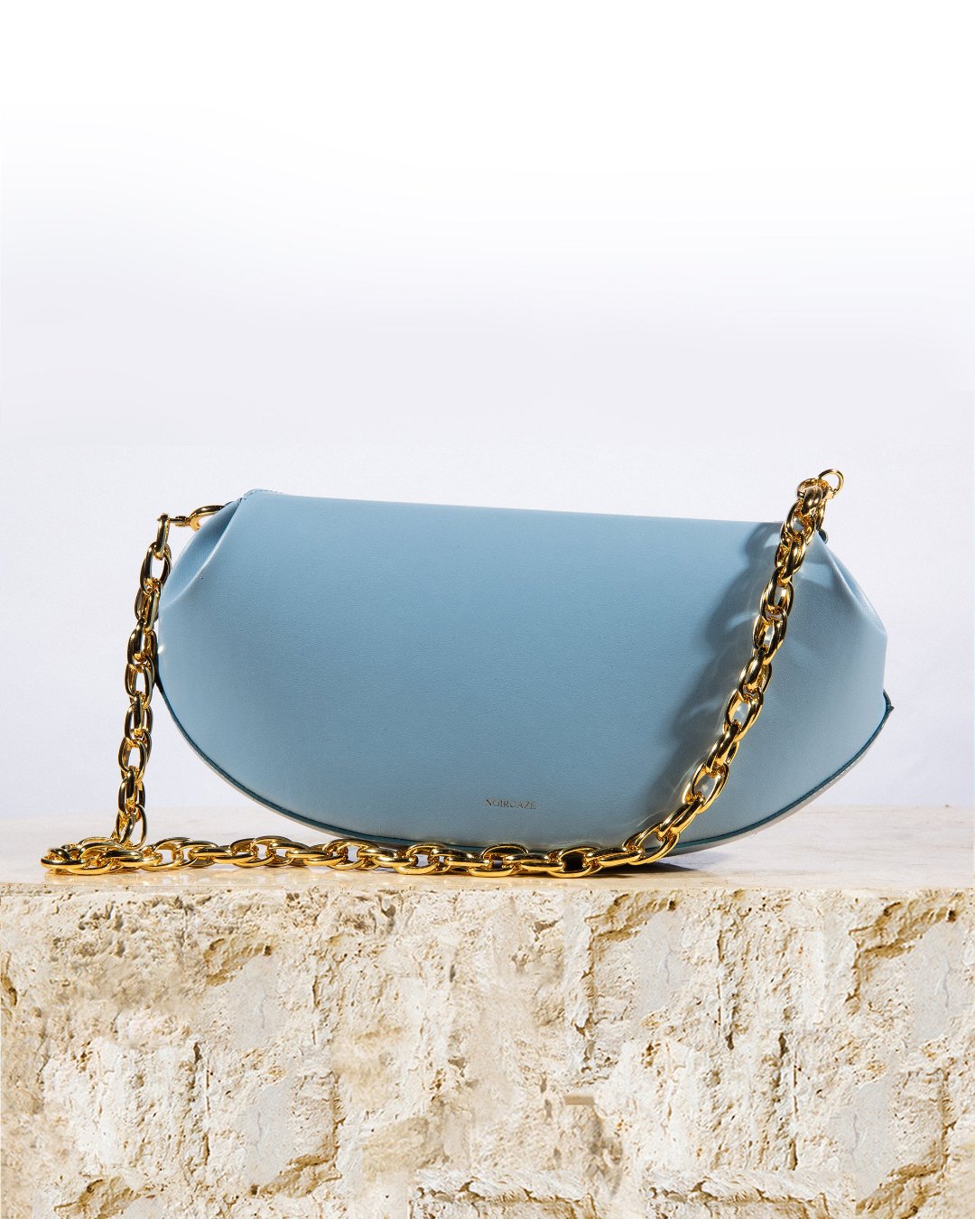 Bond Bag in Baby blue shown from a three-quarter angle highlighting its asymmetrical silhouette and gold chain strap