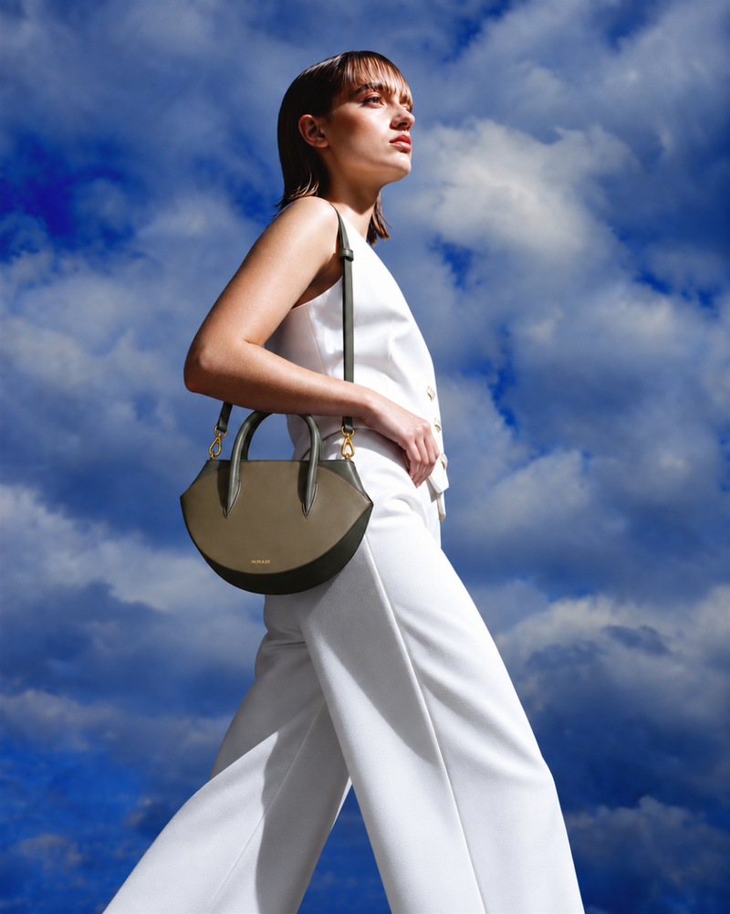 A model in a white sleeveless top and wide-leg trousers carries a structured olive green leather handbag with gold-tone hardware against a dramatic blue sky with white clouds.