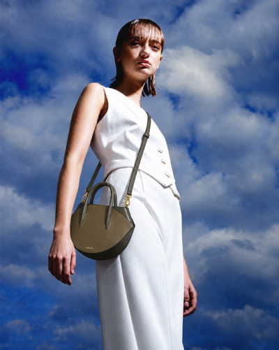 A model in a white sleeveless dress carries a structured olive green leather handbag with gold-tone hardware against a dramatic blue sky with white clouds.