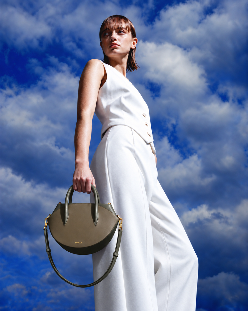 A model in a white sleeveless jumpsuit holds a structured olive green handbag with dark green handles against a dramatic blue sky with white clouds.