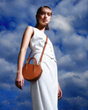 A model in a white sleeveless dress carries a brown leather crescent-shaped shoulder bag with gold-tone hardware against a dramatic blue sky with clouds.