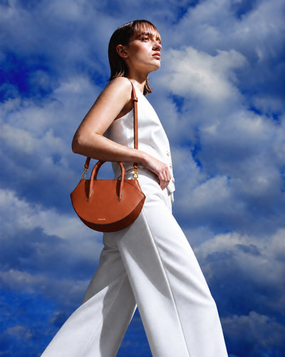 A woman in a white sleeveless top and wide-leg trousers carries a brown leather crescent-shaped shoulder bag with gold hardware against a dramatic blue sky with white clouds.