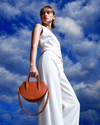A model in a white sleeveless jumpsuit holds a brown leather crescent-shaped handbag against a dramatic blue sky with white clouds.