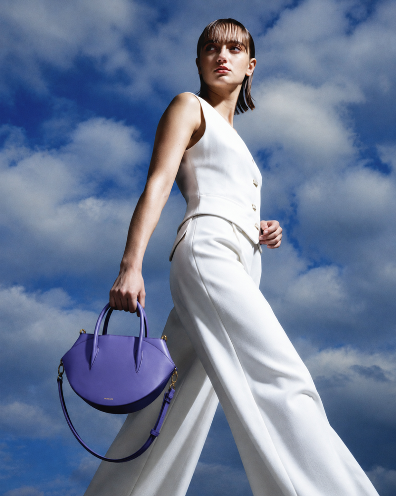 A model in a white sleeveless vest and wide-leg trousers carries a purple leather top-handle handbag against a blue sky with clouds.
