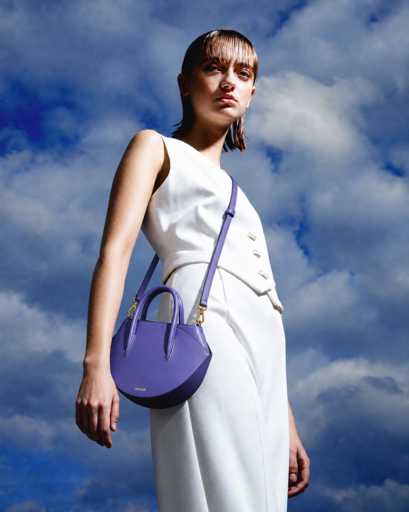 A model wearing a white sleeveless dress carries a purple leather top-handle shoulder bag with gold-tone hardware against a dramatic blue sky with clouds.