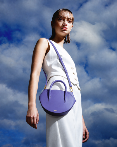 A model wearing a white sleeveless dress stands against a dramatic blue sky with clouds, carrying a structured purple leather handbag with gold-tone hardware.