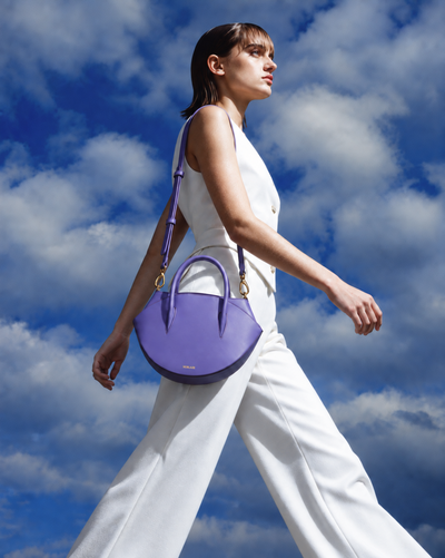 A woman in a white sleeveless top and white wide-leg pants walks outdoors against a blue sky with clouds, carrying a purple crescent-shaped shoulder bag with gold-tone hardware.